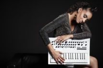 Fashionable woman posing with a synthesizer against a dark background.